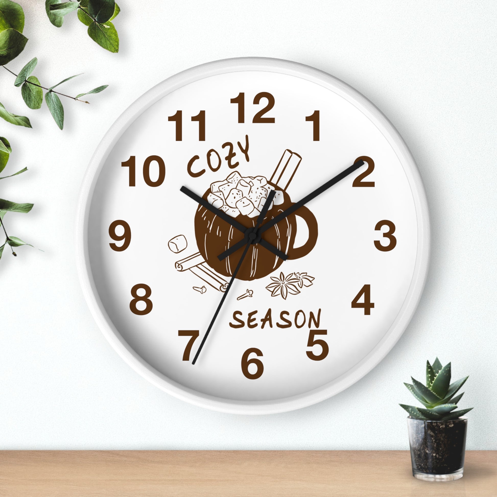 A round white wall clock with a brown mug and cinnamon stick illustration in the center, surrounded by the words 'COZY SEASON' and brown numerals. The clock is mounted on a white wall above a light wood surface, with small green plants in the corners.