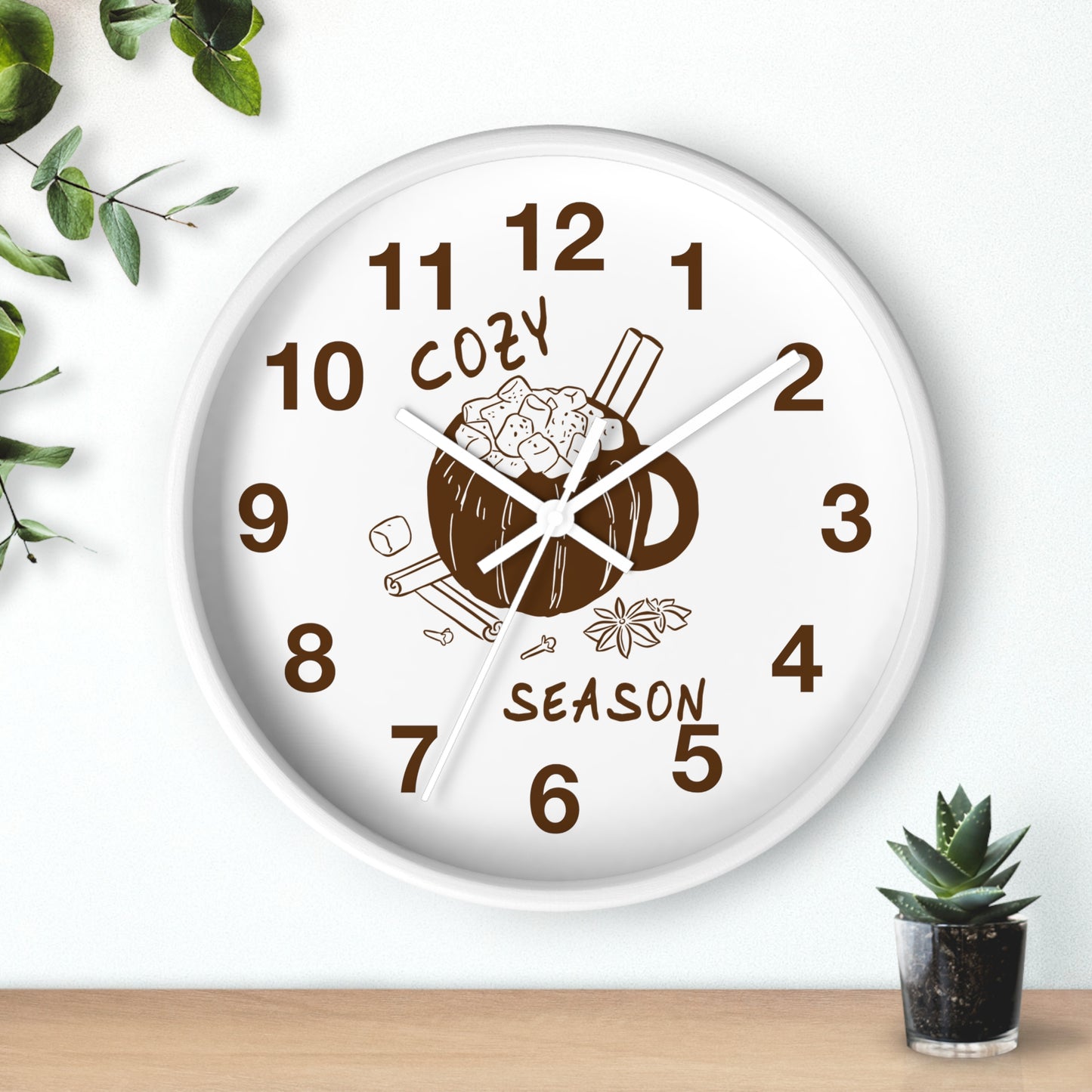 A round white wall clock with a brown mug and cinnamon illustration on the face, with the words 'COZY SEASON' and brown numerals. The clock is mounted on a white wall above a light wood surface, with small green plants in the corners.
