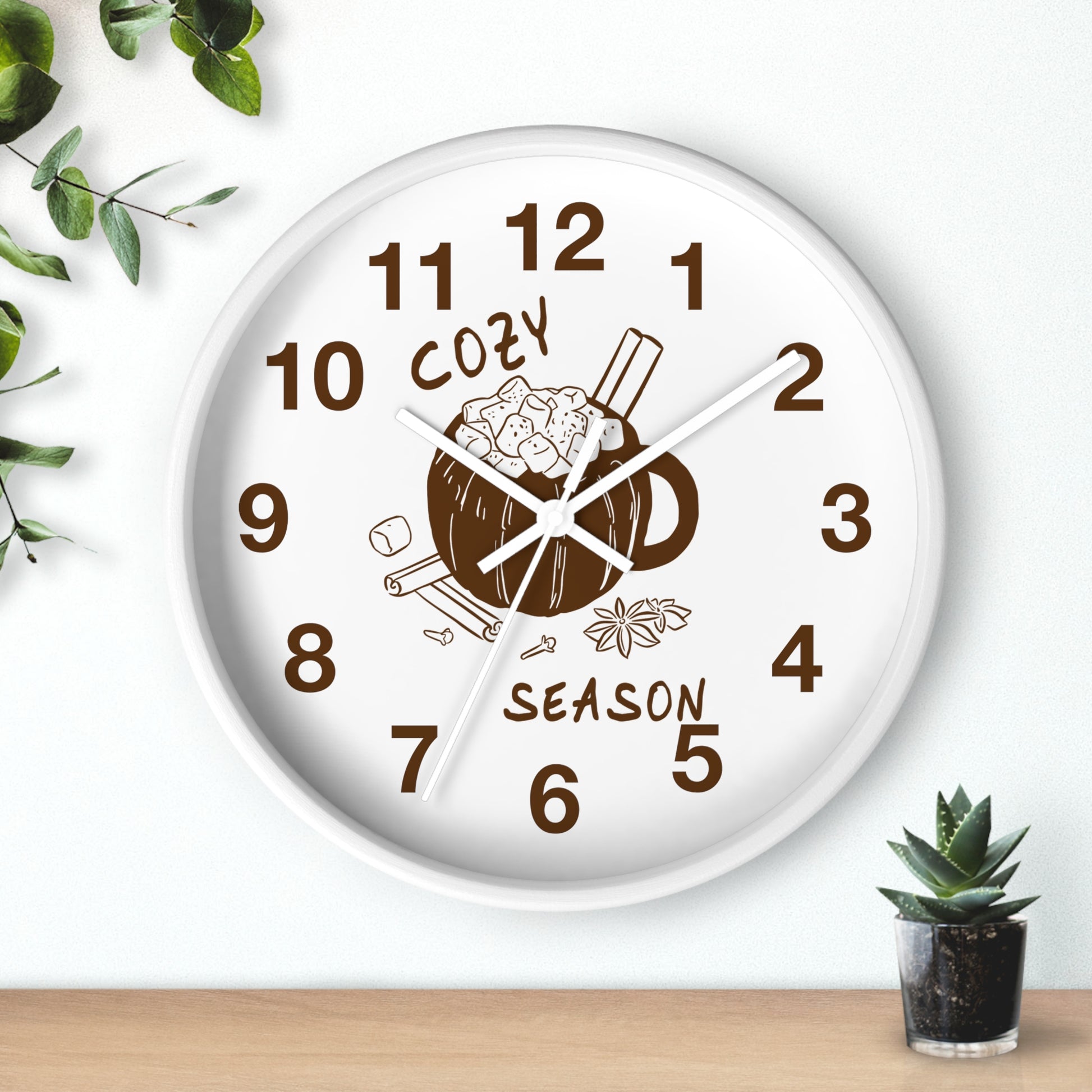 A round white wall clock with a brown mug and cinnamon illustration on the face, with the words 'COZY SEASON' and brown numerals. The clock is mounted on a white wall above a light wood surface, with small green plants in the corners.