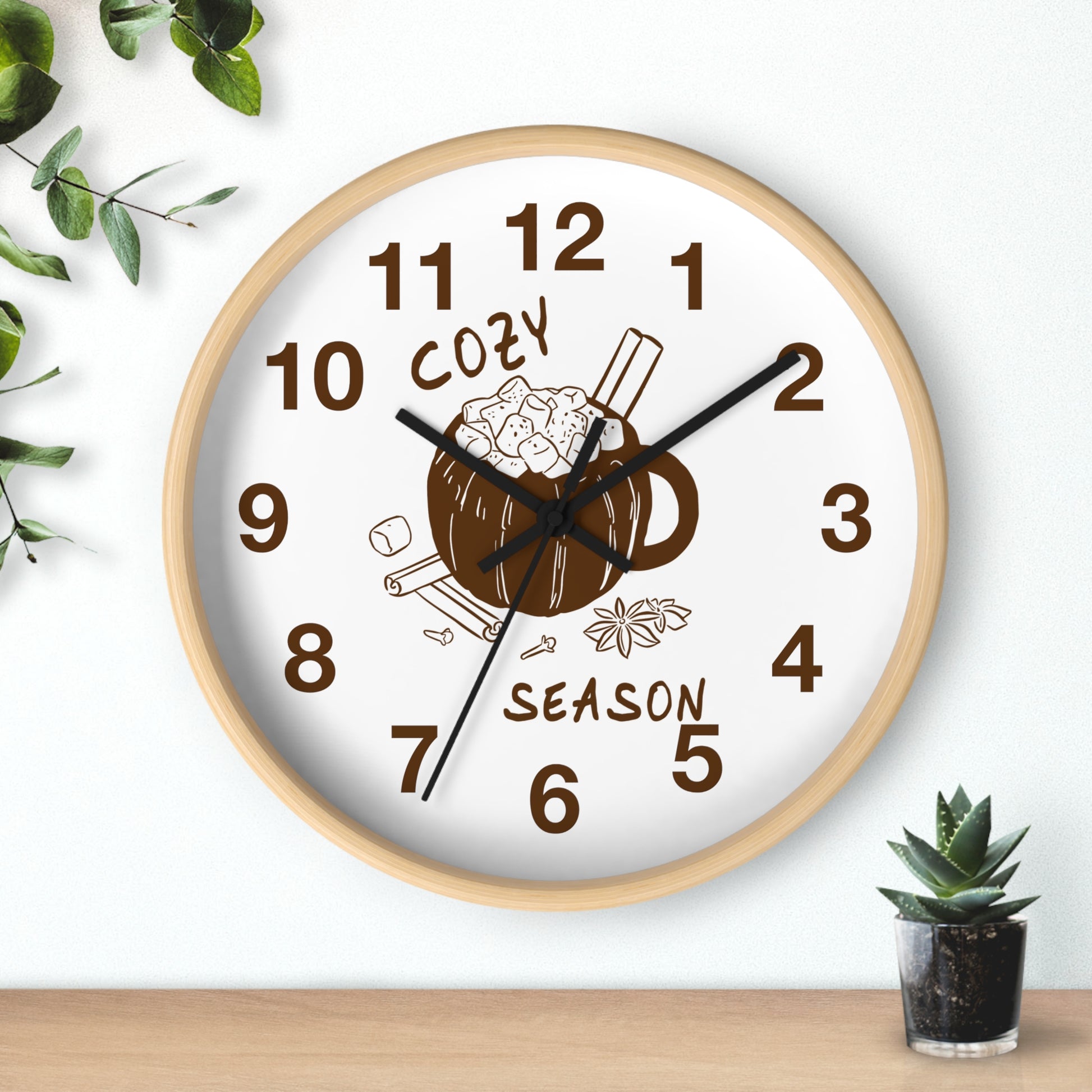 A round wall clock with a light wood frame and a white face, featuring a cozy mug illustration with cinnamon sticks and the words 'COZY SEASON' in brown. The clock is mounted on a white wall above a wooden surface, with small green plants in the corners.