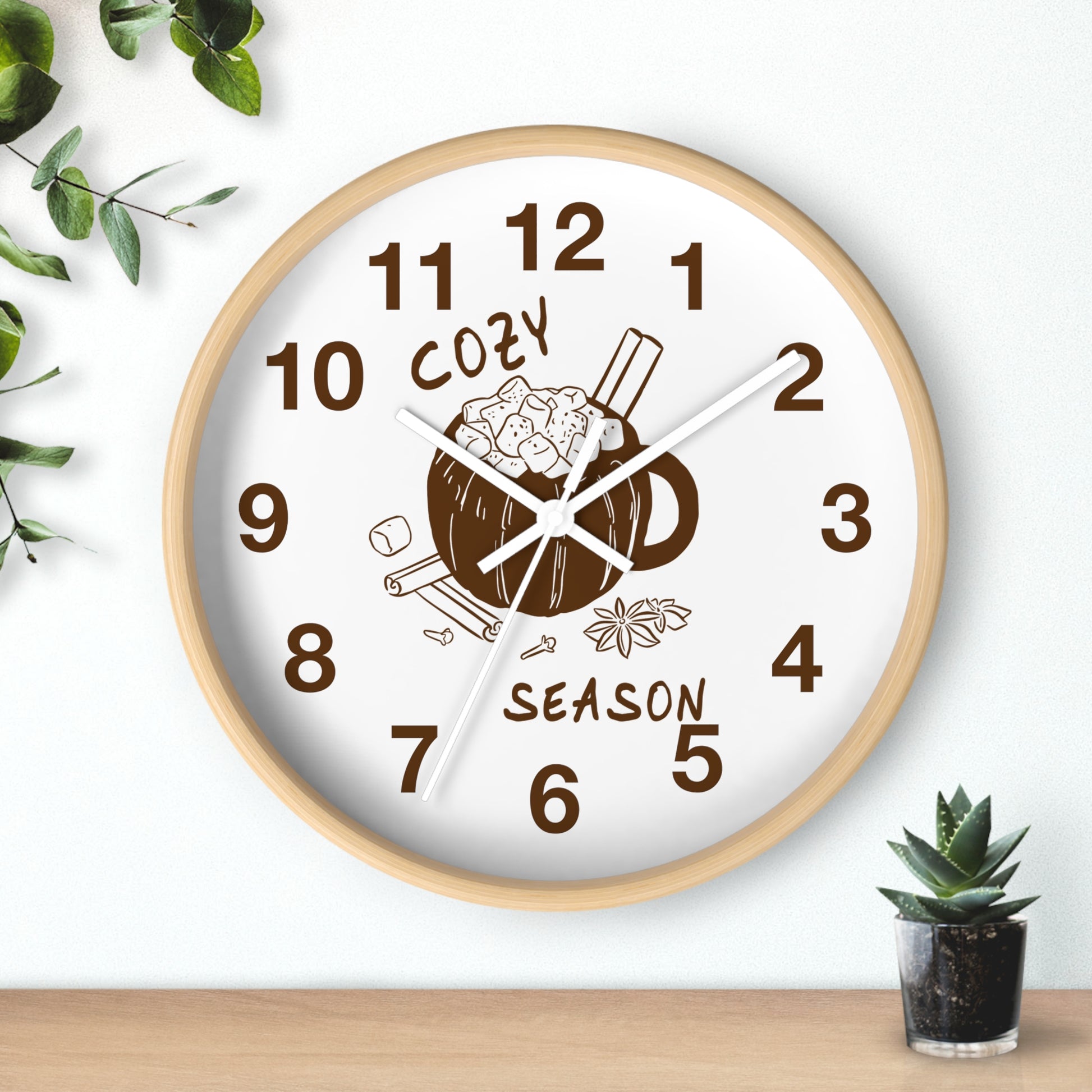 A round wall clock with a light wood frame and a white face, featuring a brown mug illustration with cinnamon sticks and the words 'COZY SEASON' in brown. The clock is shown hanging on a white wall above a wooden surface, with small green plants in the corners.