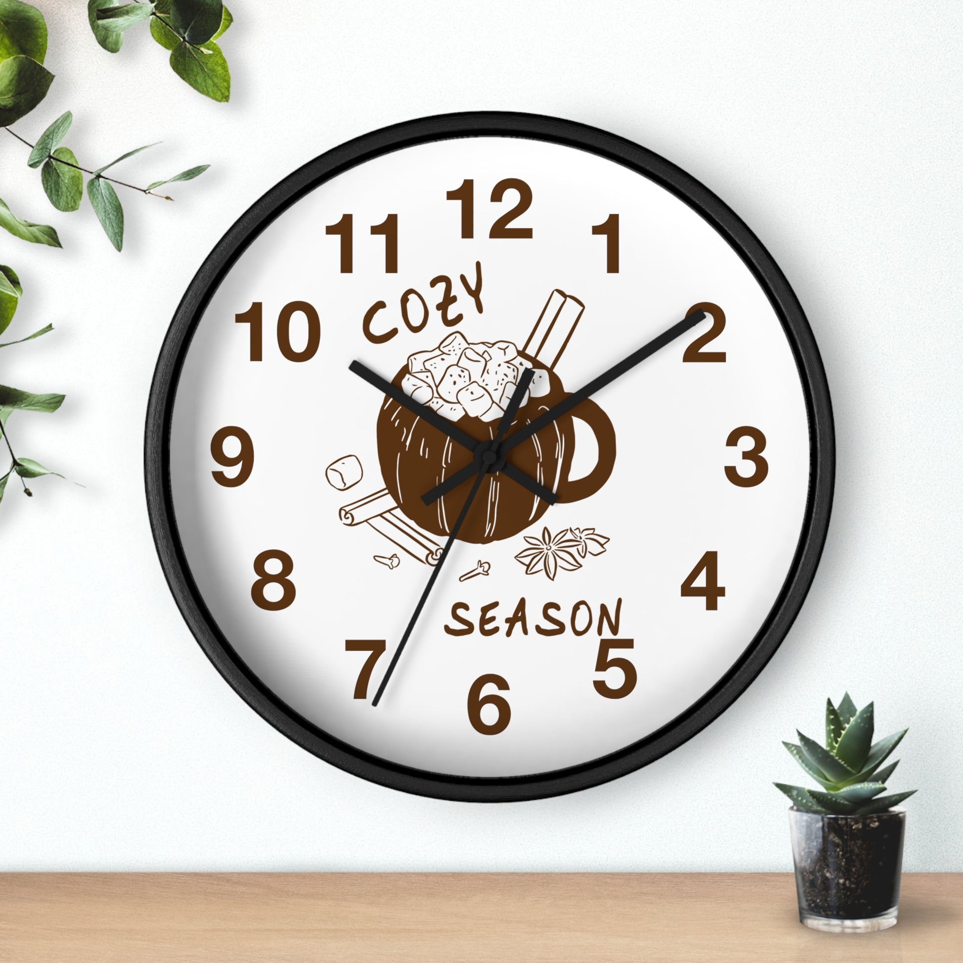 A round wall clock with a black frame and a white face, featuring a brown mug illustration with cinnamon sticks and the words 'COZY SEASON' in brown. The clock is mounted on a white wall above a light wood surface, with small green plants in the corners.
