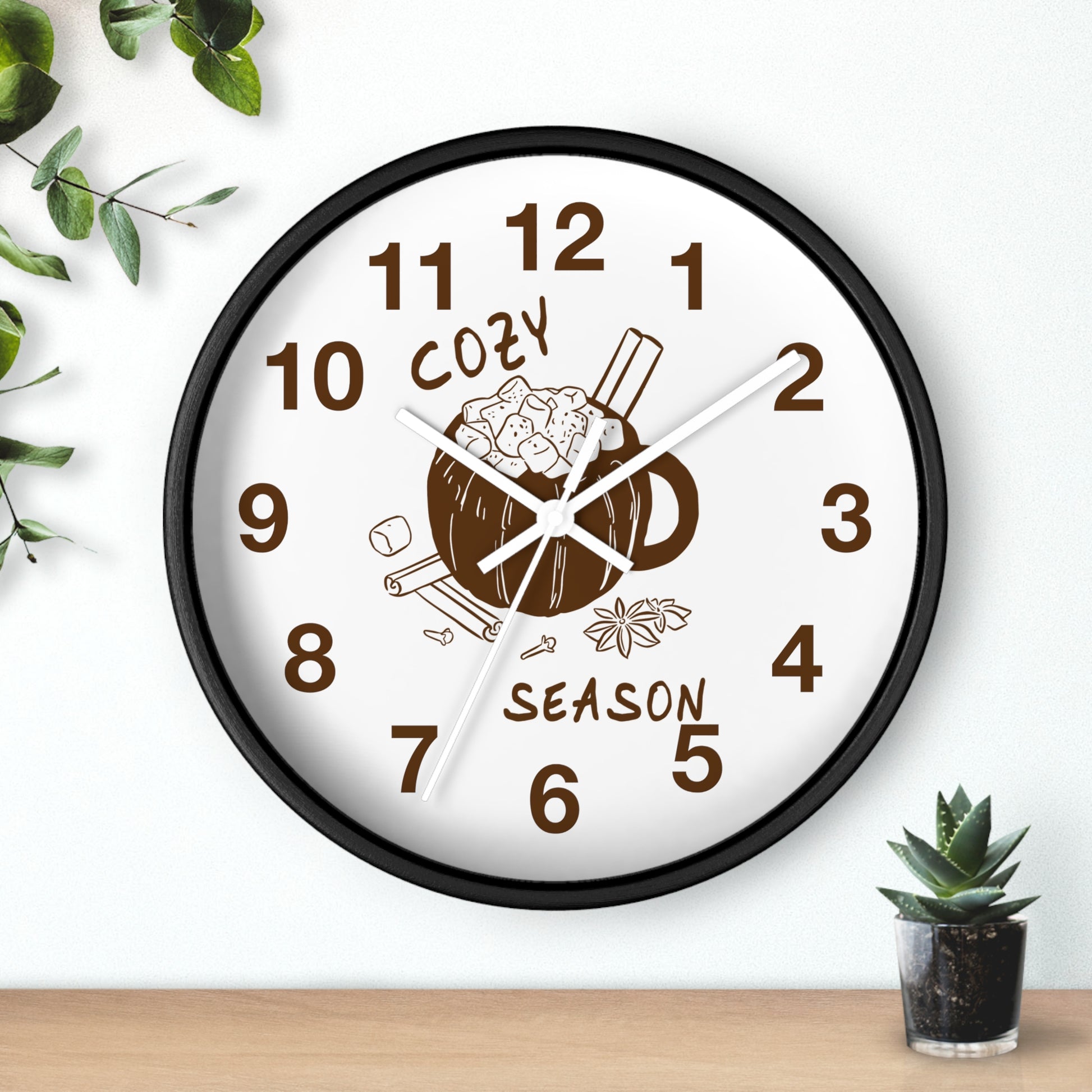 A round wall clock with a black frame and a white face, featuring a brown coffee mug illustration with cinnamon sticks and the words 'COZY SEASON' on the dial. The clock is mounted on a white wall above a light wood surface, with small green plants in the corners.