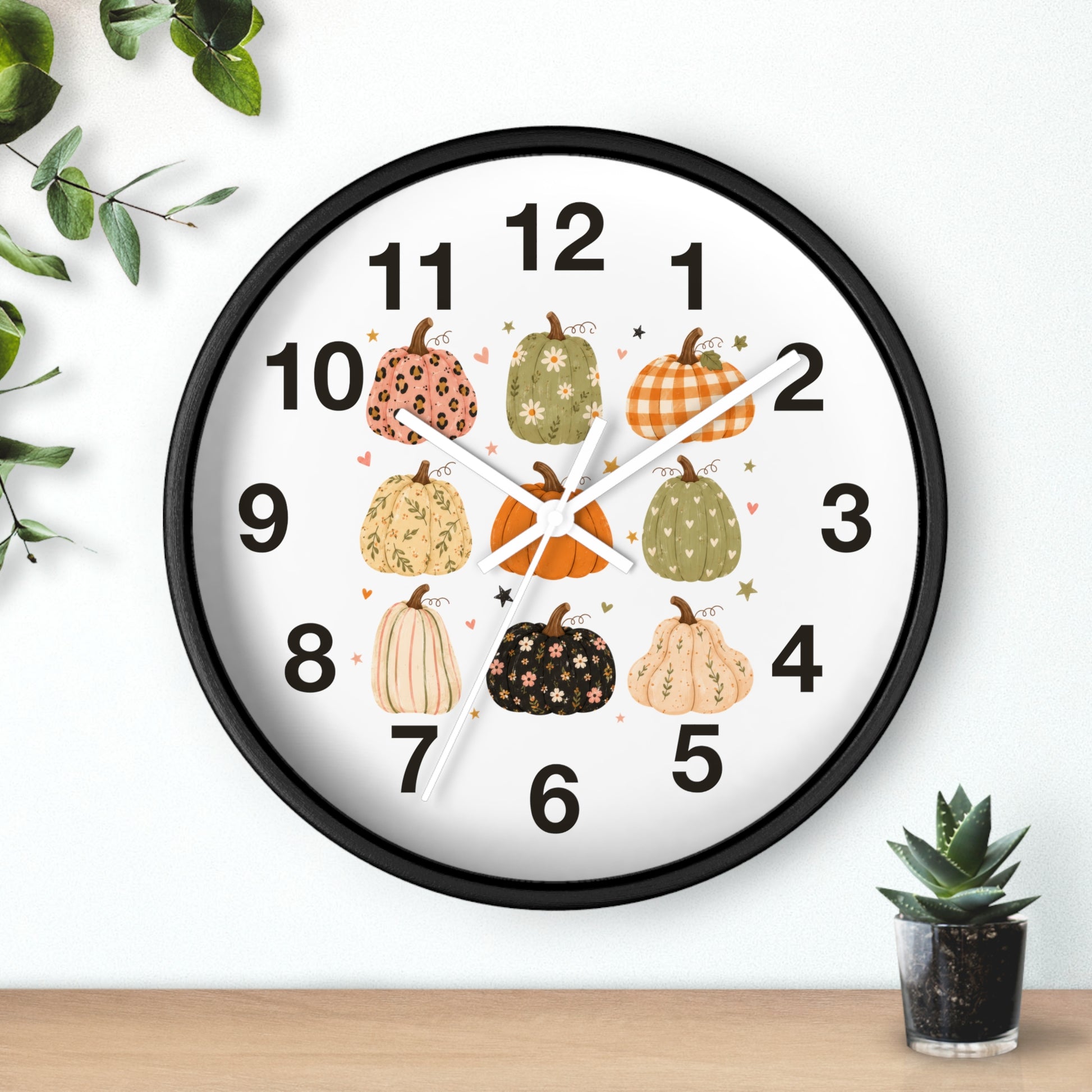 A round wall clock with a black frame and a white face, decorated with illustrated pumpkins in autumn colors around the numbers. The clock is shown hanging on a white wall above a wooden surface, with small green plants in the corners.