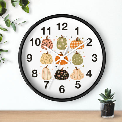 A round wall clock with a black frame and a white face, decorated with illustrated pumpkins in autumn colors around the numbers. The clock is shown hanging on a white wall above a wooden surface, with small green plants in the corners.