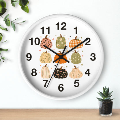 A round white wall clock with a black rim and black numerals, decorated with illustrated pumpkins in autumn colors on the face. The clock is shown hanging on a white wall above a light wood surface, with small green plants at the corners.