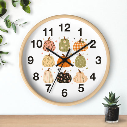 A round wall clock with a light wood frame and a white face, decorated with illustrated pumpkins in autumn colors and black numerals. The clock is shown hanging on a white wall above a wooden surface, with small green plants in the corners.