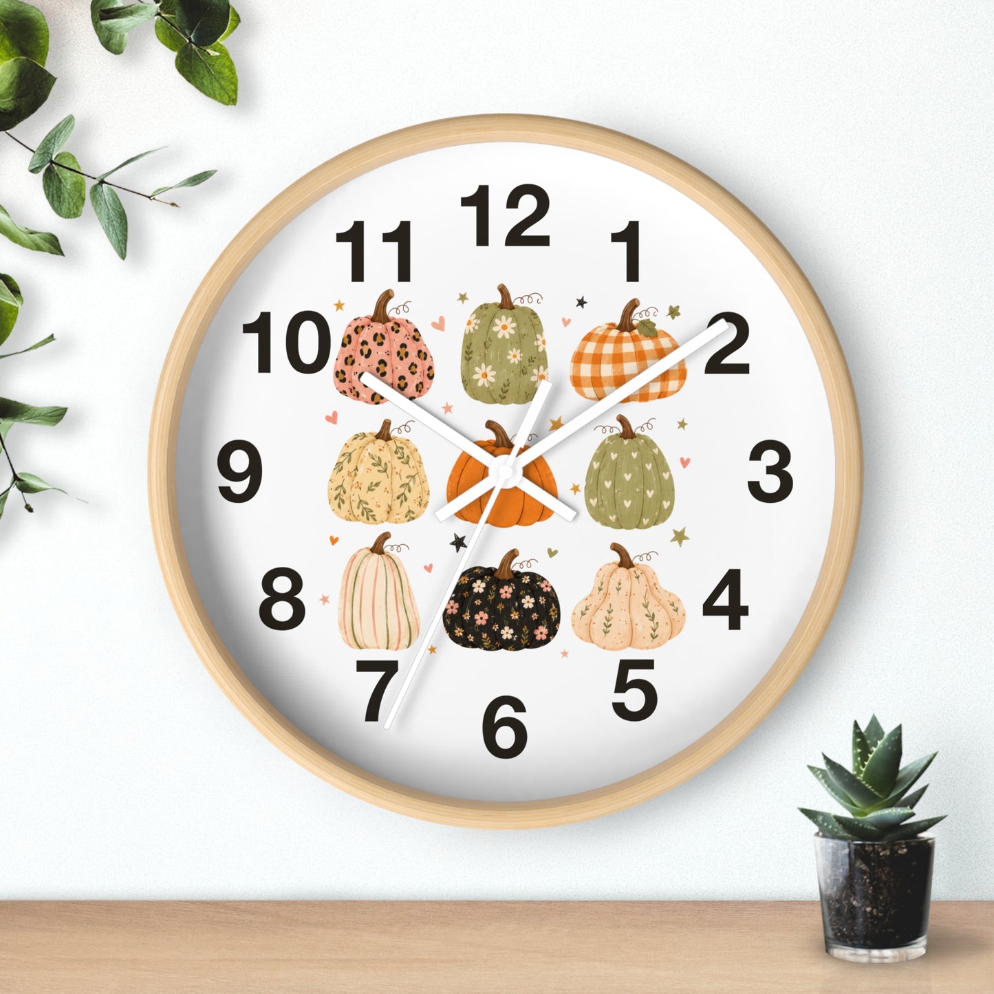 A round wall clock with a light wood frame and a white face, decorated with illustrated pumpkins in autumn colors and black numerals. The clock is shown hanging on a white wall above a wooden surface, with small green plants in the corners.