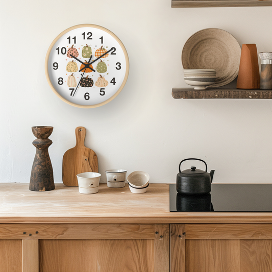 A round wall clock with a light wood frame and a white face featuring colorful autumn leaves and black Arabic numerals, styled in a warm kitchen setting above a wooden counter with ceramic dishes and a black kettle.
