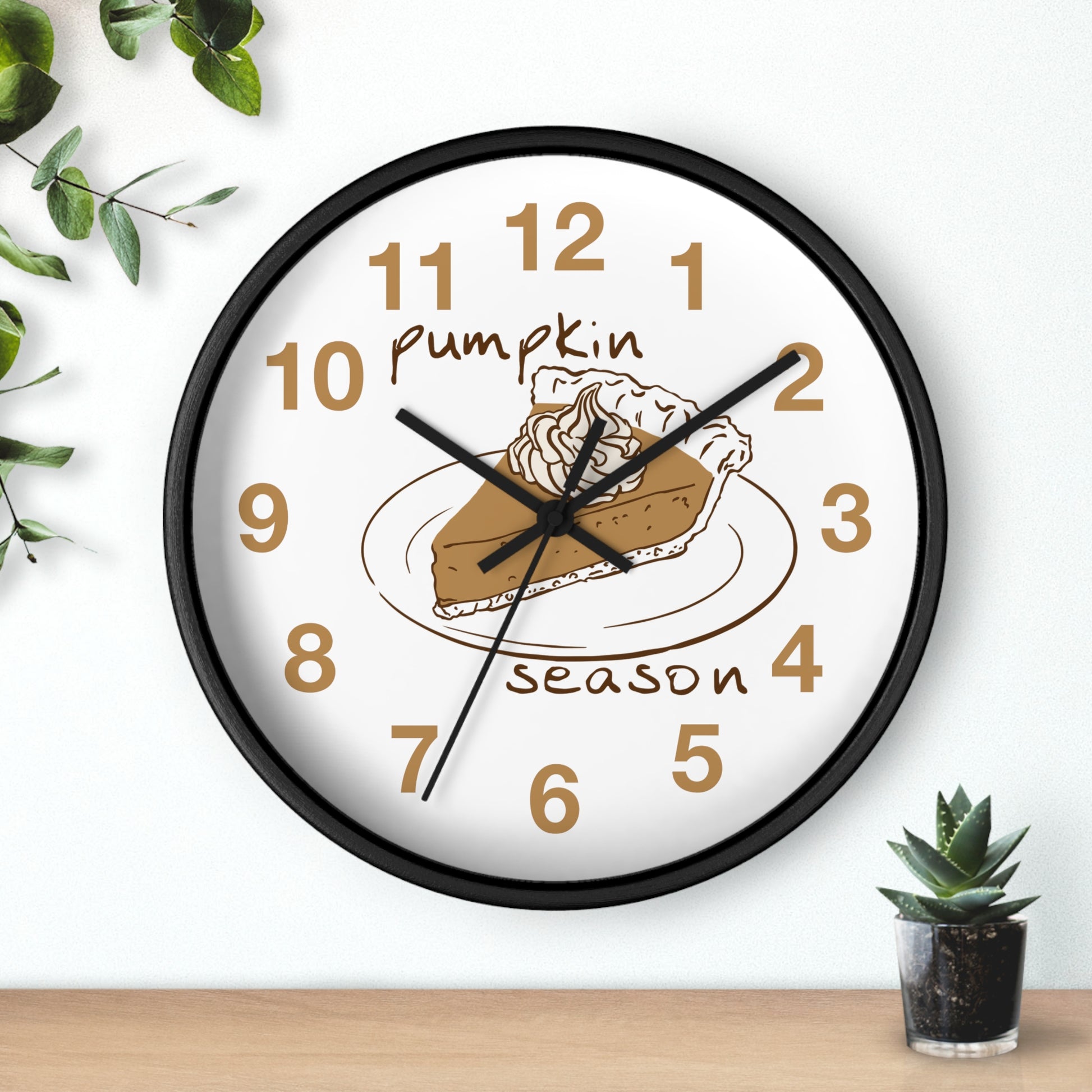 A round wall clock with a black frame and a white face, featuring a pumpkin pie slice illustration and the words 'Pumpkin Season' in brown. The clock is shown hanging on a white wall above a wooden surface, with small green plants in the corners.