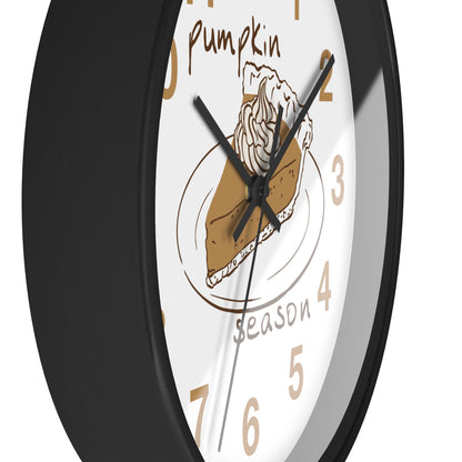 A close-up of a round wall clock with a black frame and a white face featuring a pumpkin pie illustration, tan numerals, and the words 'pumpkin season'.