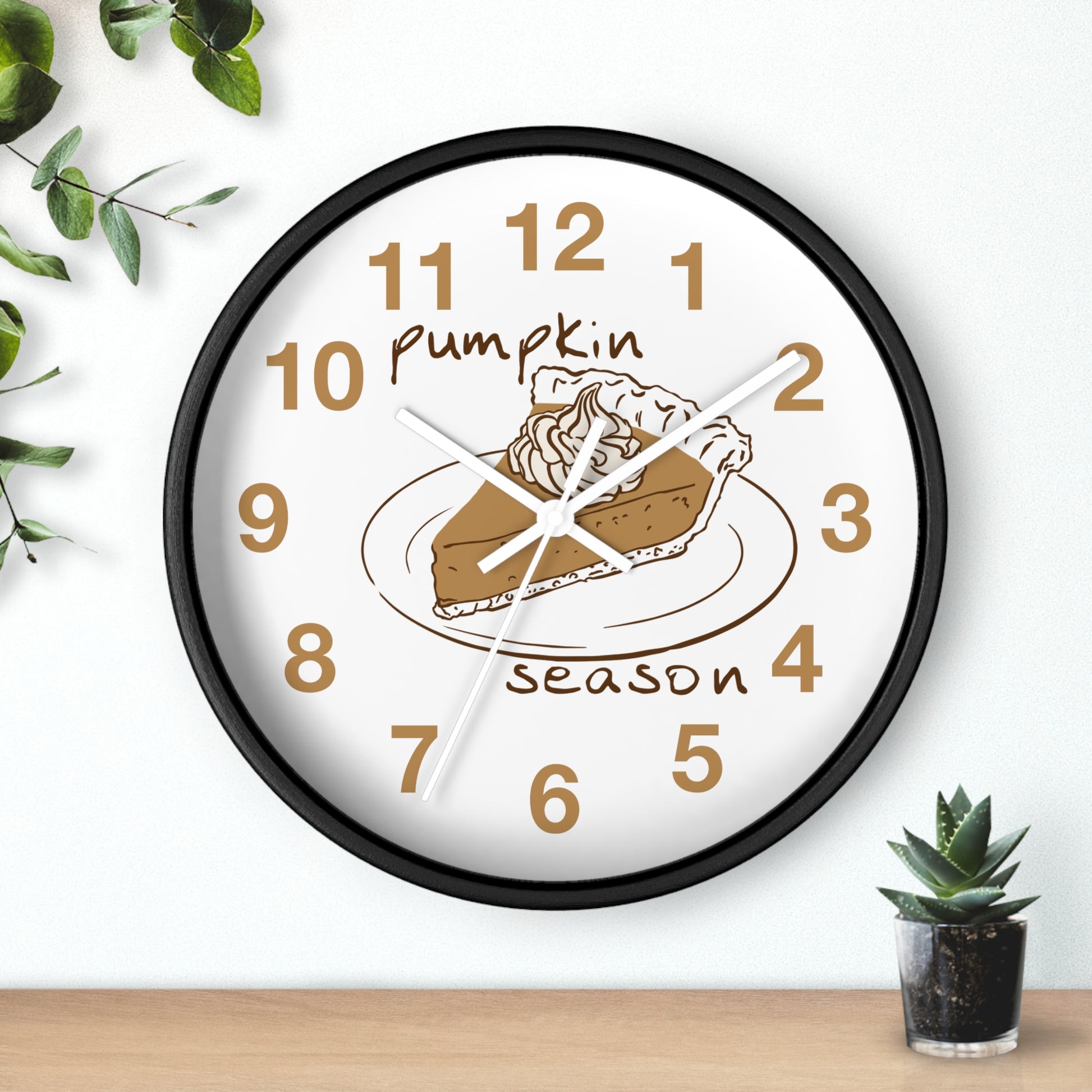 A round wall clock with a black frame and a white face, featuring a pumpkin pie illustration and the words 'Pumpkin Season' in brown. The clock is shown hanging on a white wall above a wooden surface, with decorative green leaves and a small succulent nearby.