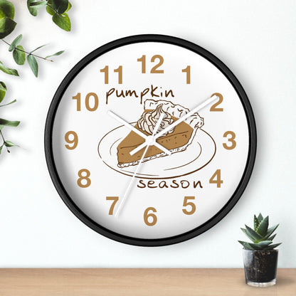 A round wall clock with a black frame and a white face, featuring a pumpkin pie illustration and the words 'Pumpkin Season' in brown. The clock is shown hanging on a white wall above a wooden surface, with decorative green leaves and a small succulent nearby.