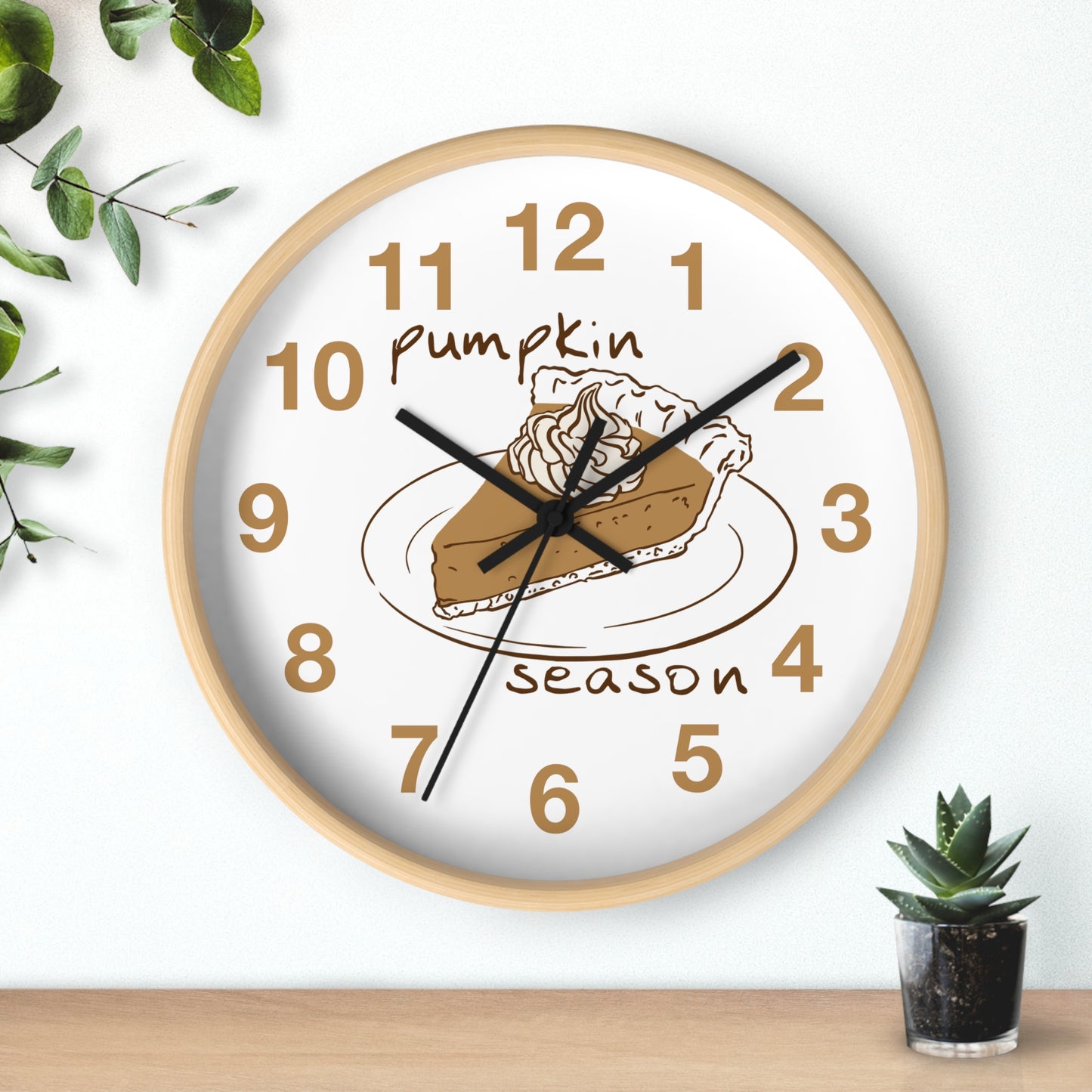 A round wall clock with a light wood frame and a white face, featuring a pumpkin pie illustration and the words 'Pumpkin season' in brown. The clock is mounted on a white wall above a wooden surface, with decorative green leaves and a small succulent nearby.
