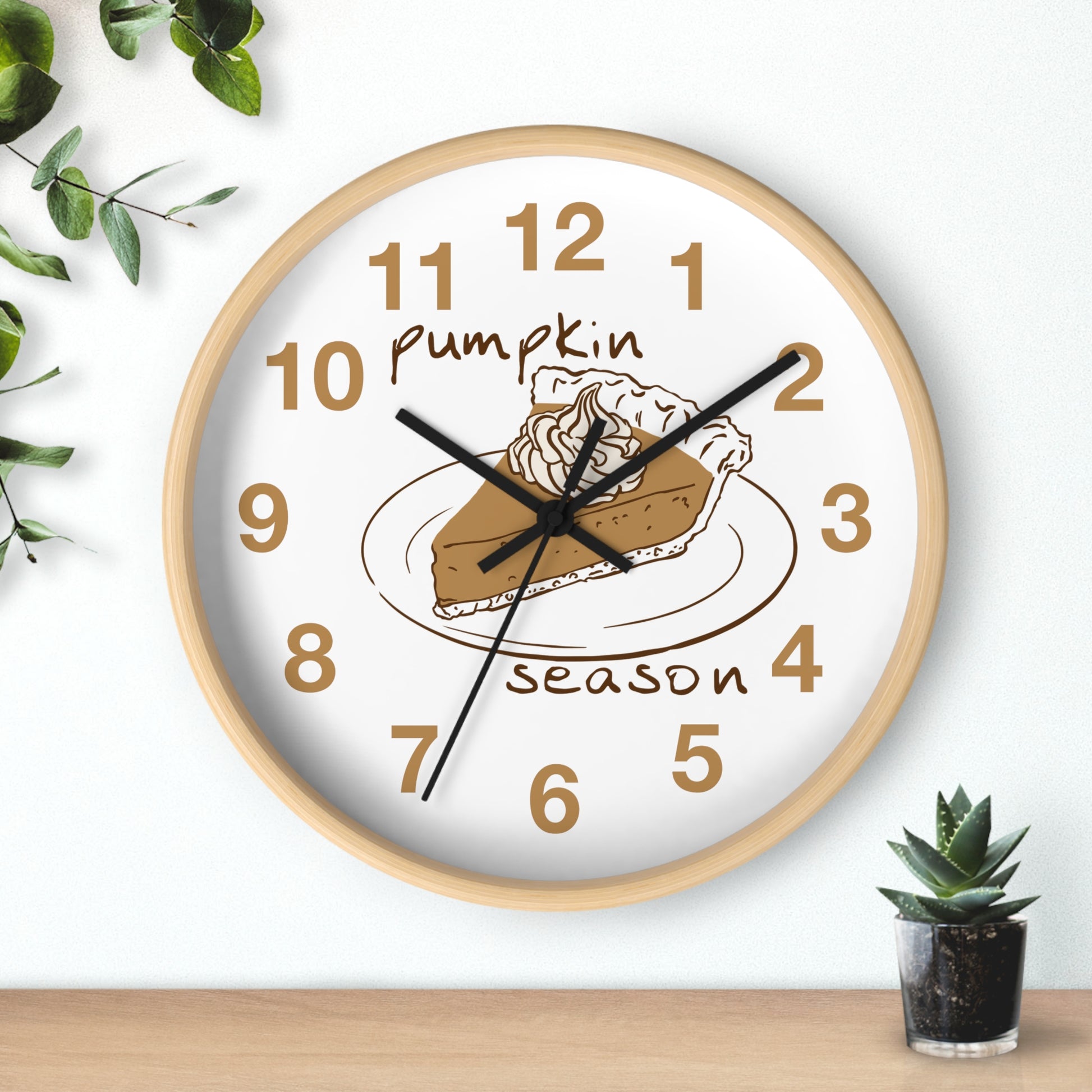 A round wall clock with a light wood frame and a white face, featuring a pumpkin pie illustration and the words 'Pumpkin season' in brown. The clock is mounted on a white wall above a wooden surface, with decorative green leaves and a small succulent nearby.