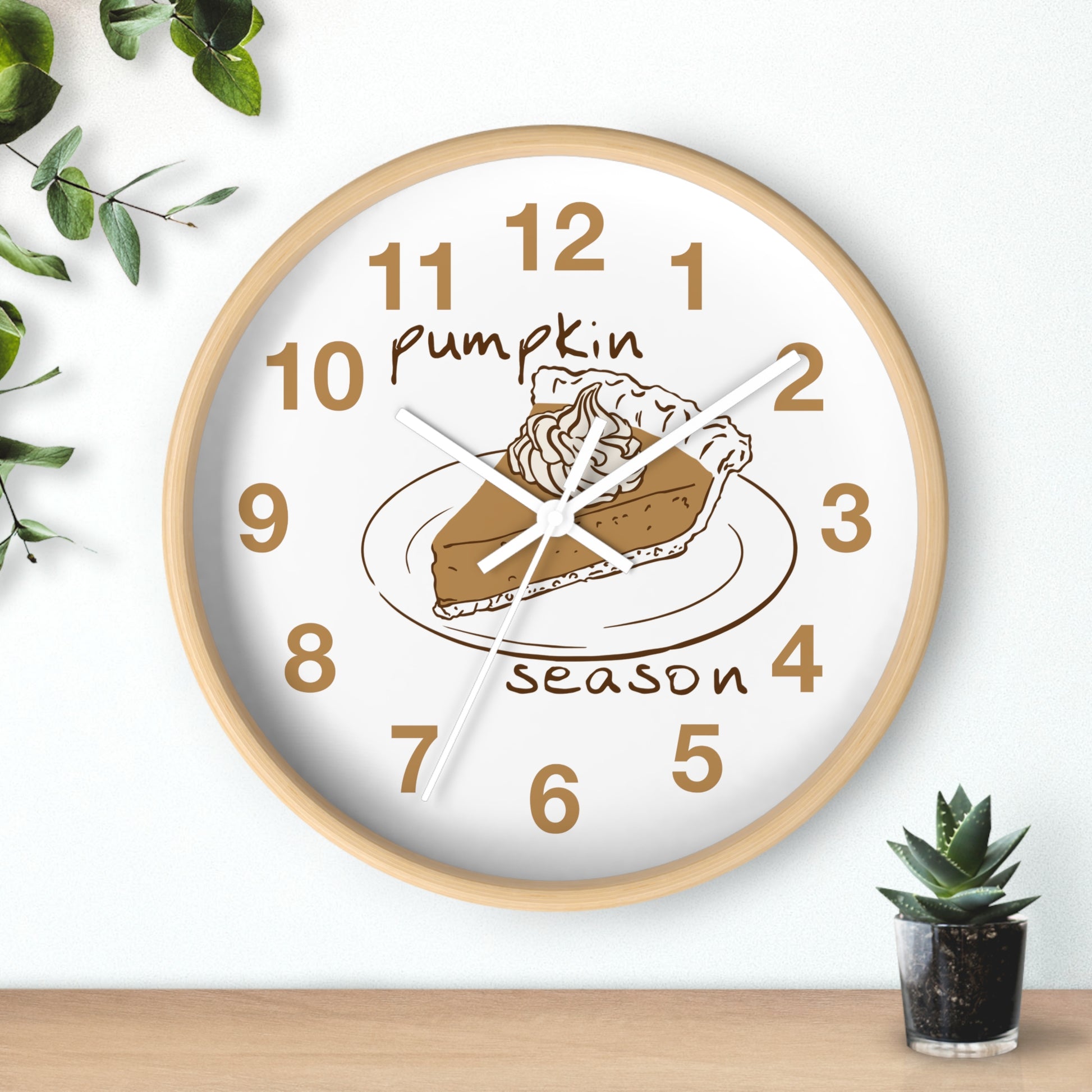 A round wall clock with a light wood frame and a white face, featuring a pumpkin pie illustration and the words 'Pumpkin Season' in brown. The clock is shown hanging on a white wall above a wooden surface, with small green plants in the corners.