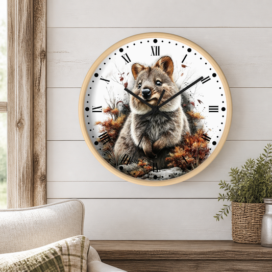 A round wall clock with a light wood frame and a printed illustration of a quokka centered on a white face with black Roman numerals, mounted on a white shiplap wall above a wooden surface with a chair and plant nearby.
