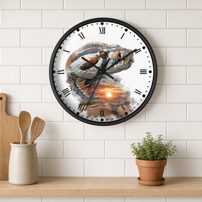 A round wall clock with a black frame and Roman numerals, featuring a detailed graphic of a coiled snake over a sunset landscape on the clock face. The clock is mounted on a white subway tile wall above a wooden shelf with kitchen utensils and a potted plant.