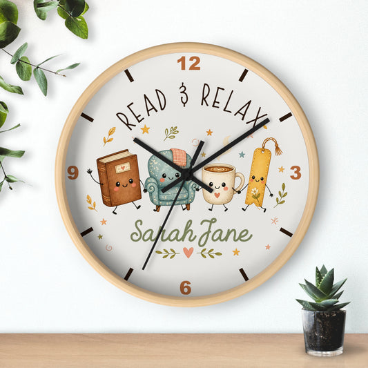 A round wall clock with a light wood frame and a white face, featuring cute illustrated characters and the words 'READ & RELAX' and the name 'Sarah Jane'. The clock is shown hanging on a white wall above a wooden surface, with decorative green leaves and a small potted succulent nearby.