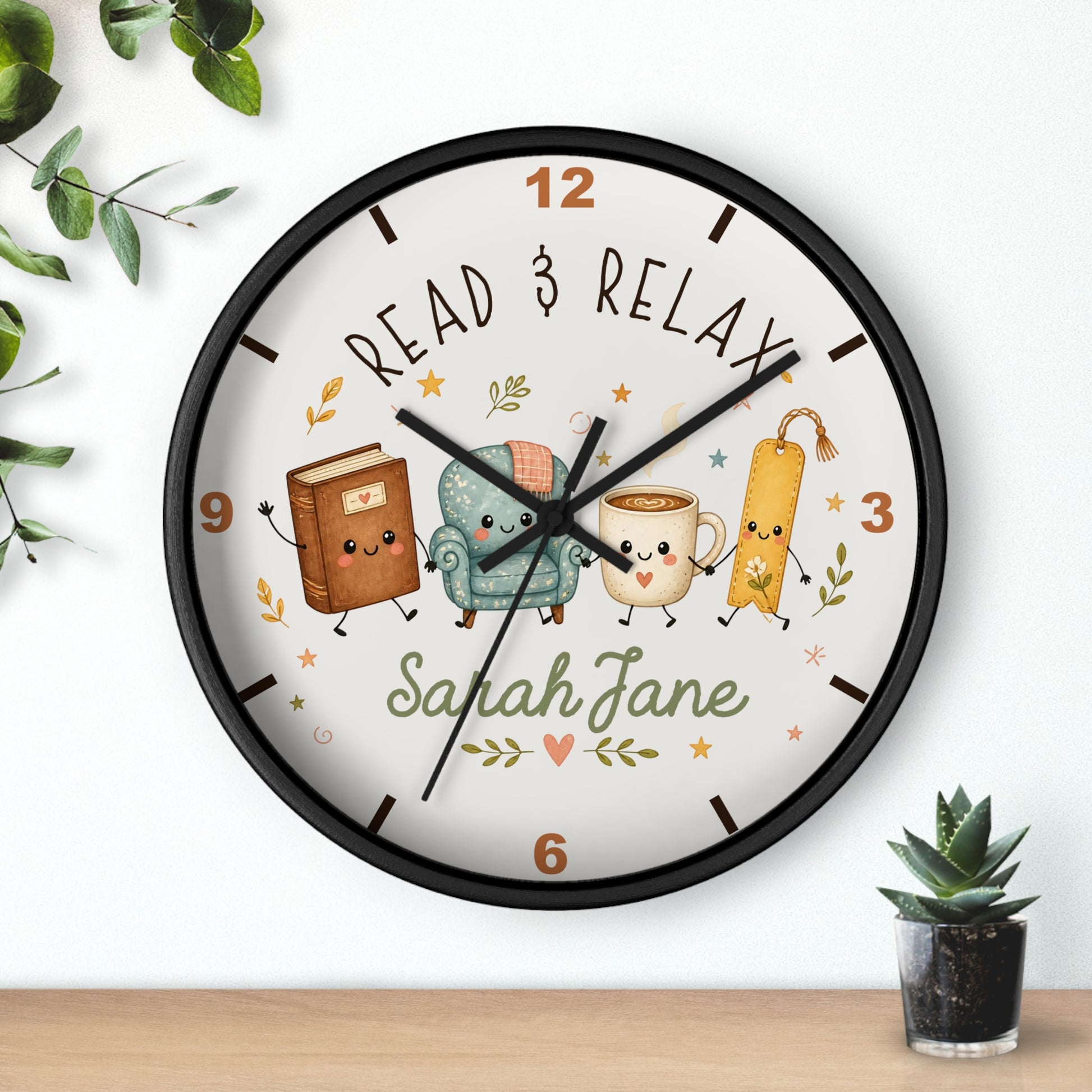 A round wall clock with a black frame and a white face, featuring cartoon illustrations of a book, armchair, mug, and bookmark, with the text 'READ & RELAX' and the name 'Sarah Jane'. The clock is shown hanging on a white wall above a wooden surface, with small green plants in the corners.