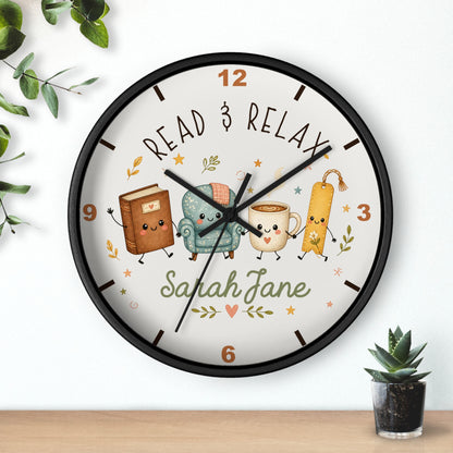 A round wall clock with a black frame and a white face, featuring cartoon illustrations of a book, armchair, mug, and bookmark, with the text 'READ & RELAX' and the name 'Sarah Jane'. The clock is shown hanging on a white wall above a wooden surface, with small green plants in the corners.