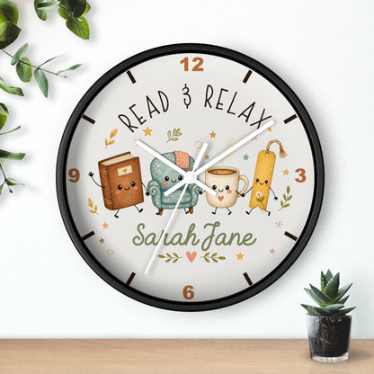 A personalized round wall clock with a black frame and a whimsical reading-themed dial. The clock face features cute illustrations of a book, a cozy armchair, a smiling mug, and a bookmark, surrounded by the words 'READ & RELAX' and the custom name 'Sarah Jane'.