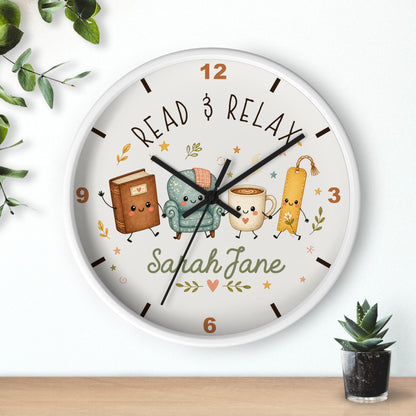 A round wall clock with a white frame and a whimsical book-themed dial. The clock face features the text 'READ & RELAX' and the personalized name 'Sarah Jane' in green script, surrounded by cute illustrations of a brown book, a teal armchair, a yellow mug, and a bookmark.