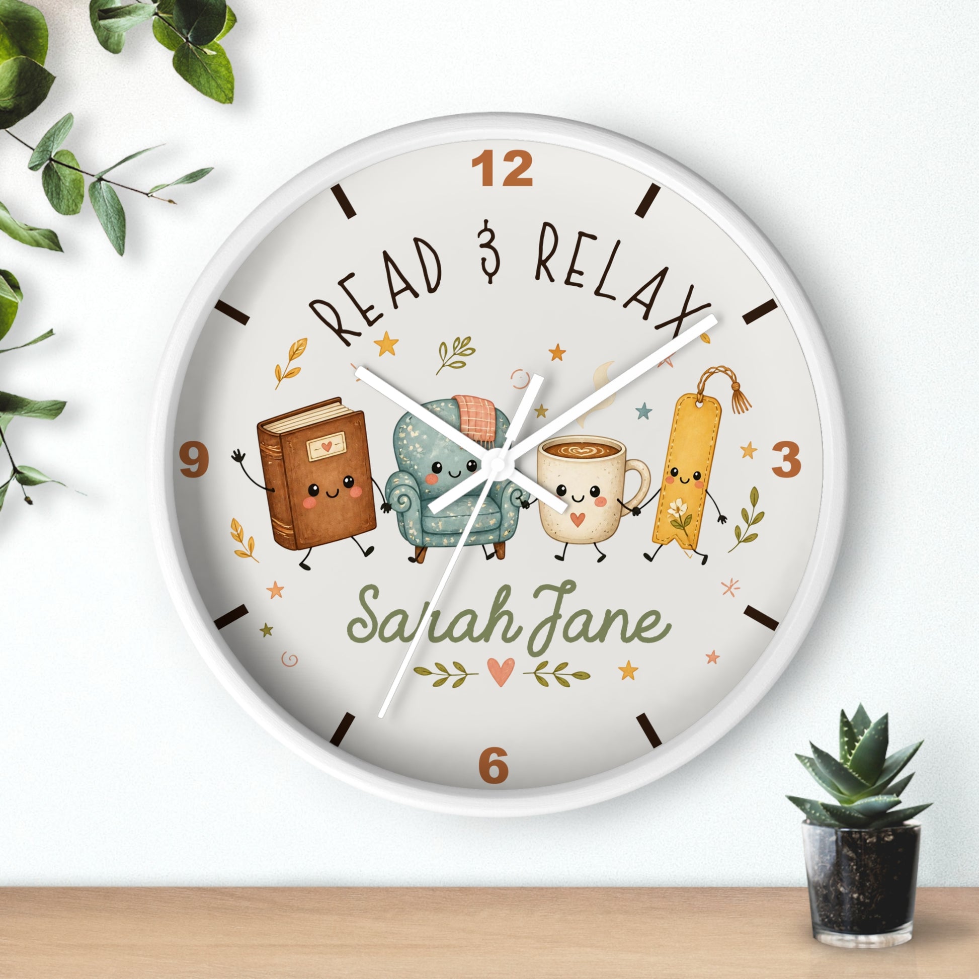 A round wall clock with a white frame and a whimsical reading-themed dial. The dial features the words 'READ & RELAX' and a custom name 'Sarah Jane' in green script, surrounded by cute illustrations of a book, a teal armchair, a mug, and a bookmark.