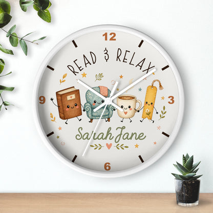 A round wall clock with a white frame and a whimsical reading-themed dial. The dial features the words 'READ & RELAX' and a custom name 'Sarah Jane' in green script, surrounded by cute illustrations of a book, a teal armchair, a mug, and a bookmark.
