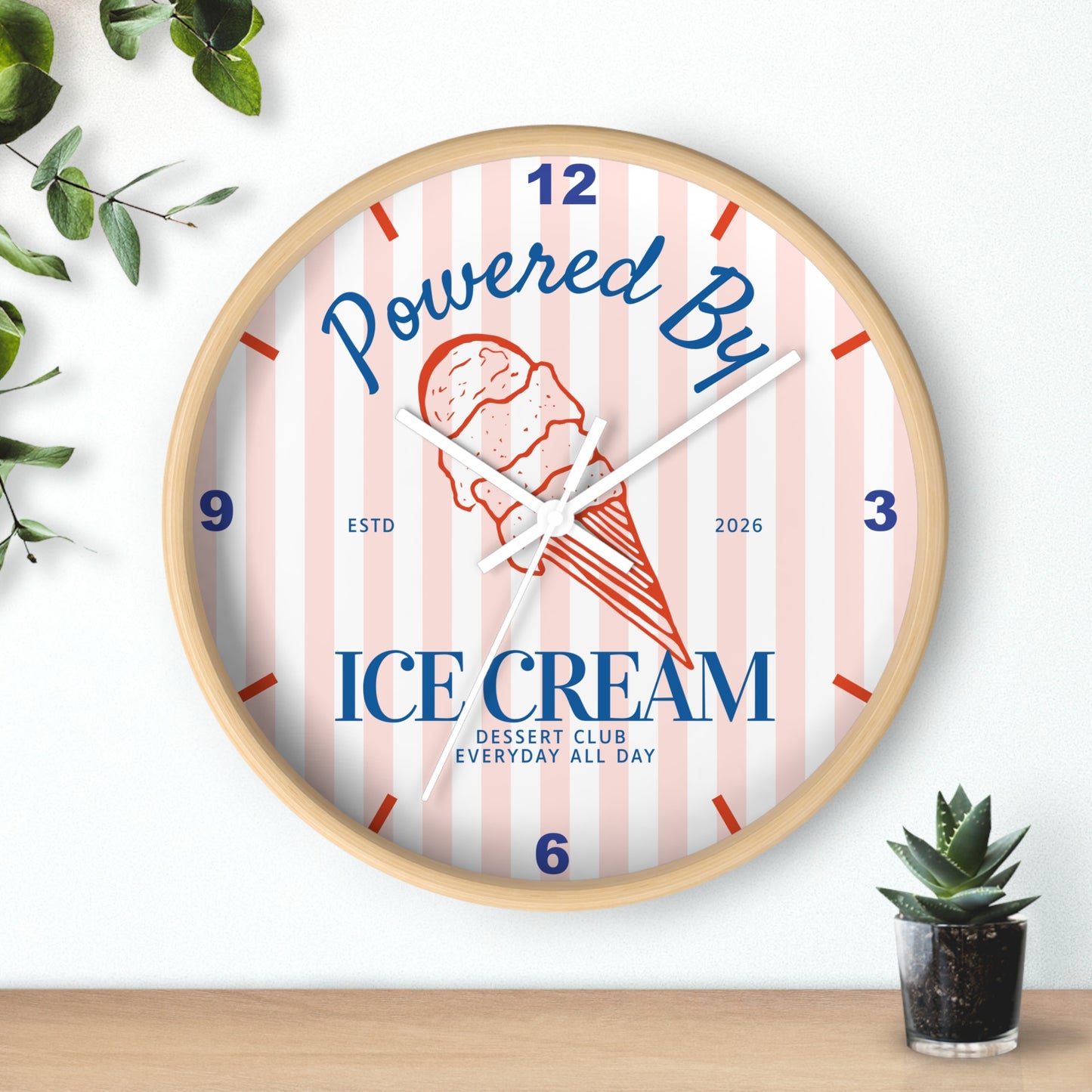 A round wall clock with a light wood frame, featuring a pink and white striped dial with blue and red text reading 'Powered By ICE cream' and a red ice cream cone illustration. The clock is styled on a wooden surface against a white wall with small green plants in the corners.
