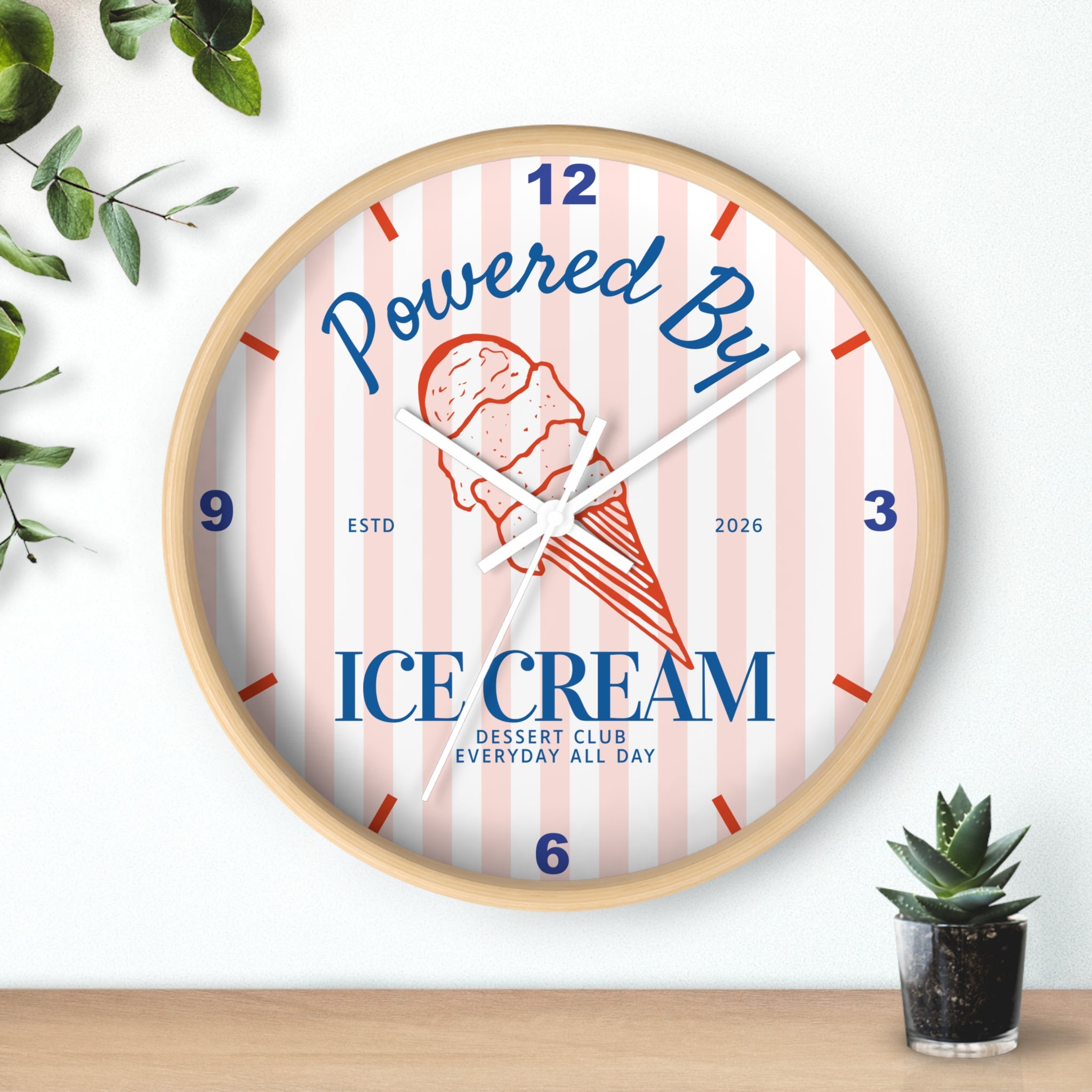 A round wall clock with a light wood frame, featuring a pink and white striped dial with blue and red text reading 'Powered By ICE cream' and a red ice cream cone illustration. The clock is styled on a wooden surface against a white wall with small green plants in the corners.