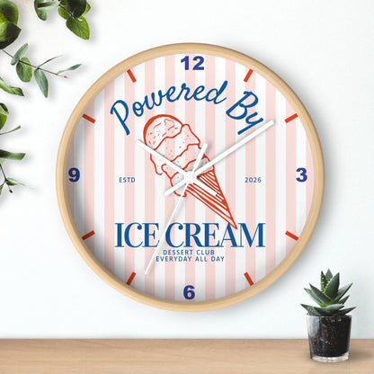 A round wall clock with a light wood frame, featuring a pink and white striped dial with blue and red text reading 'Powered By ICE cream' and a red ice cream cone illustration. The clock is styled on a wooden surface against a white wall with small green plants in the corners.