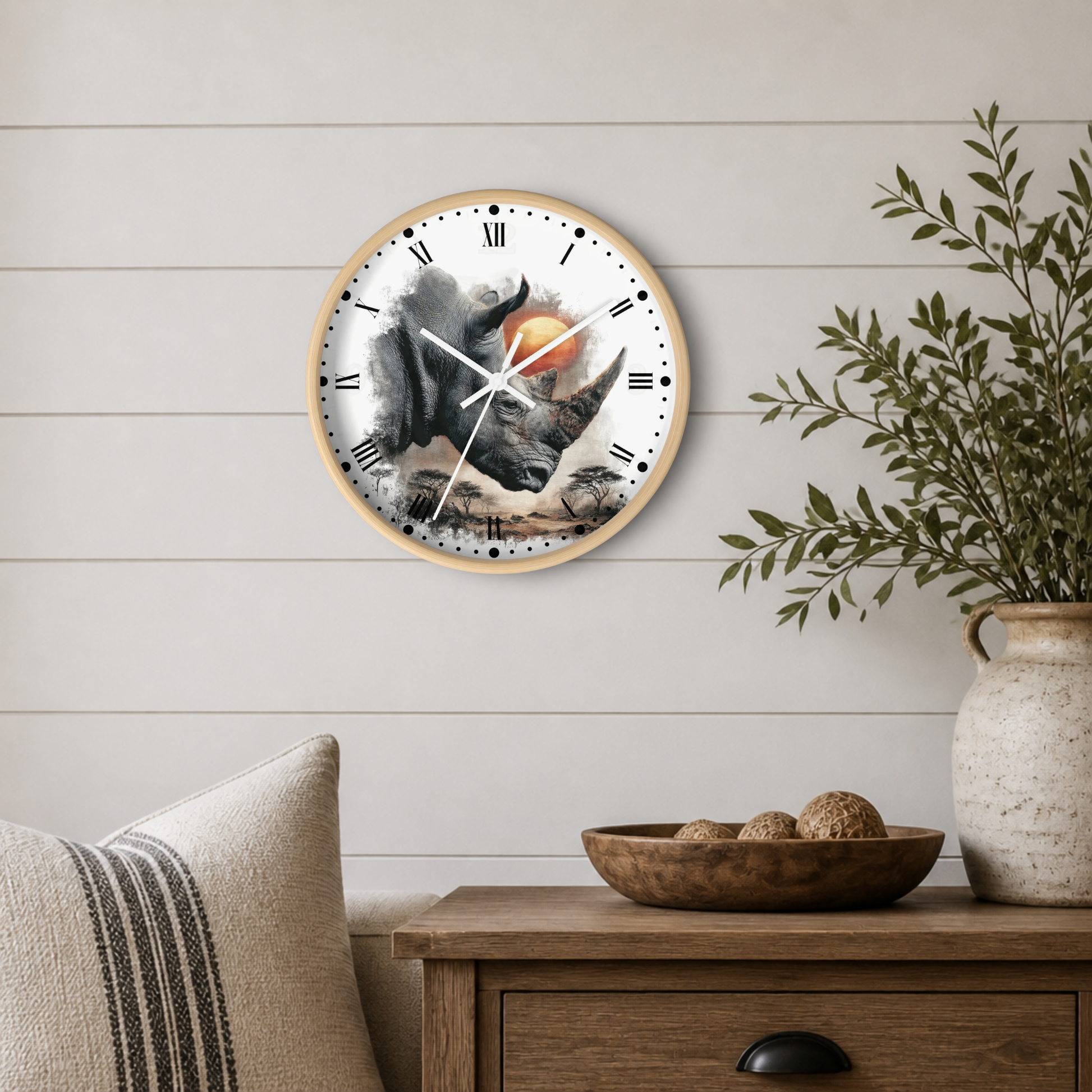 A round wall clock with a light wood frame hangs on a white shiplap wall above a wooden side table. The clock face features a detailed illustration of a gray rhinoceros in front of an orange sun, with black Roman numerals and white clock hands.