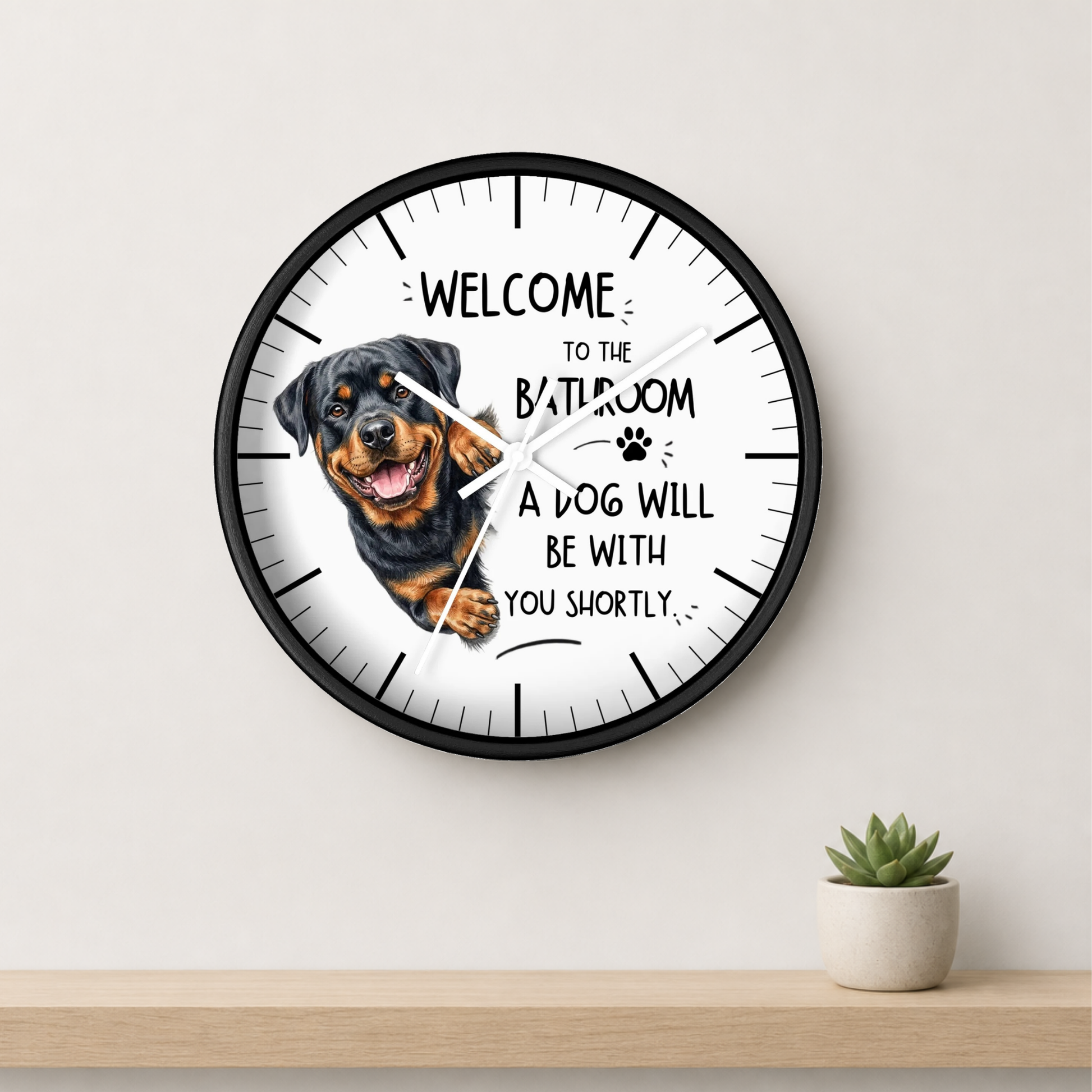 A round wall clock with a black frame and a white face, featuring a Rottweiler dog illustration and the text 'WELCOME TO THE BATHROOM A DOG WILL BE WITH YOU SHORTLY'. The clock is mounted on a light beige wall above a wooden shelf with a small potted succulent.