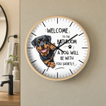 A round wall clock with a light wood frame and white face, featuring a Rottweiler illustration and the text 'WELCOME TO THE BATHROOM A DOG WILL BE WITH YOU SHORTLY.' The clock is mounted on a beige wall in a bathroom setting.