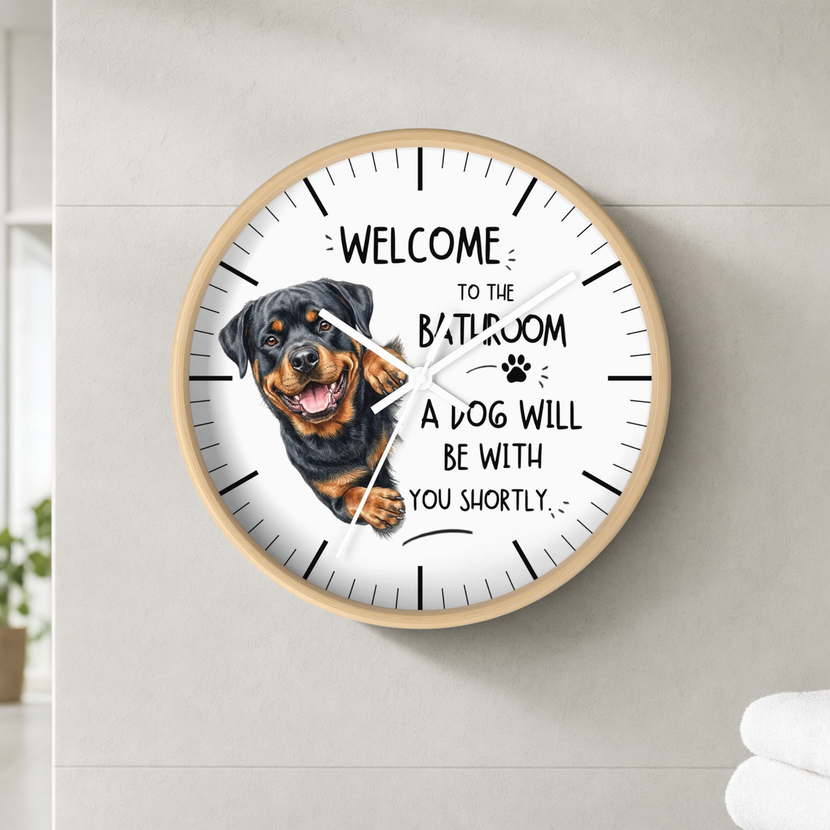 A round wall clock with a light wood frame and a white face, featuring a Rottweiler dog illustration and the text 'WELCOME TO THE BATHROOM A DOG WILL BE WITH YOU SHORTLY'. The clock is mounted on a light gray tiled wall.