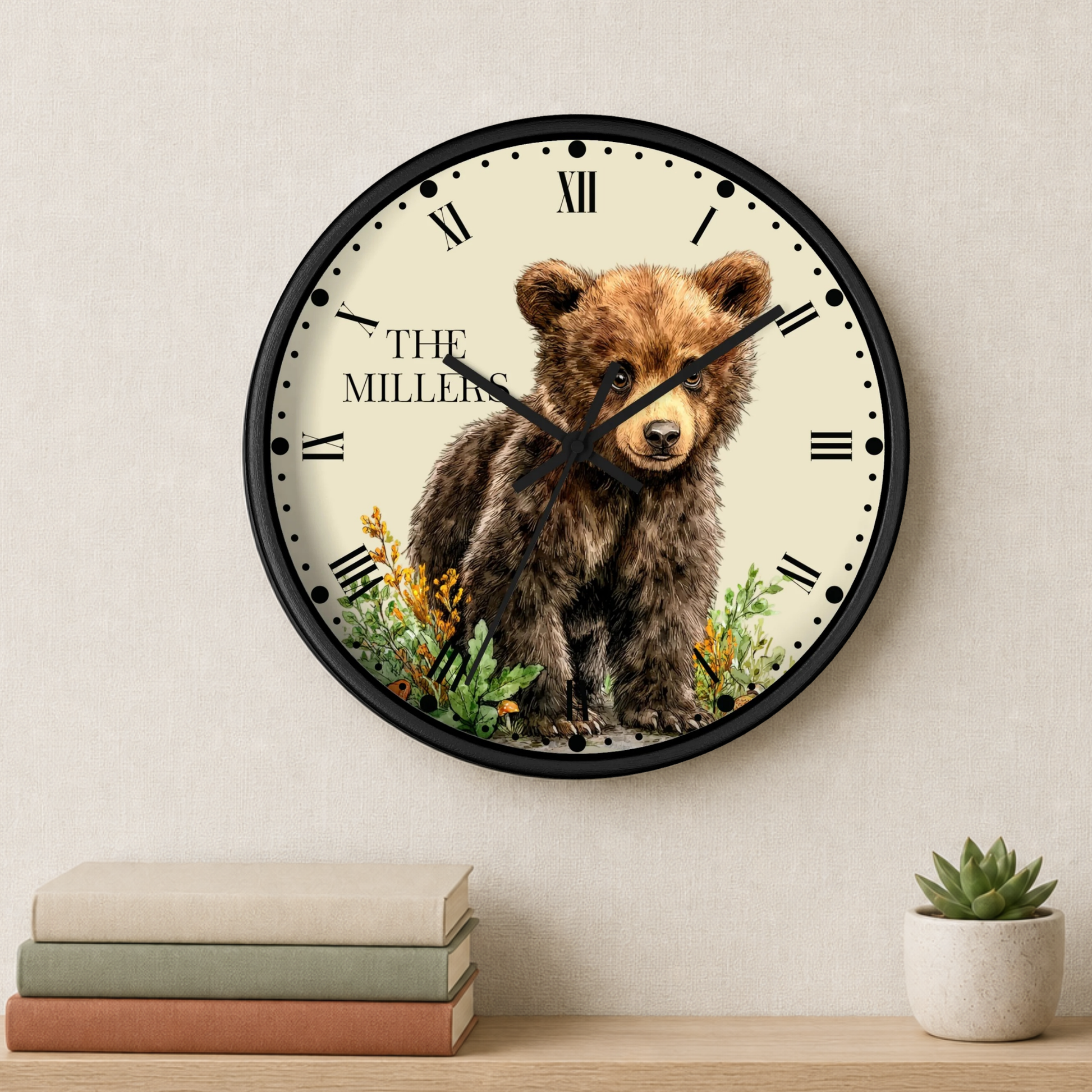 A round wall clock with a black frame and Roman numerals, featuring an illustration of a brown bear cub with the text 'THE MILLER' on the clock face. The clock is mounted on a textured beige wall above a wooden shelf with stacked books and a small potted succulent.