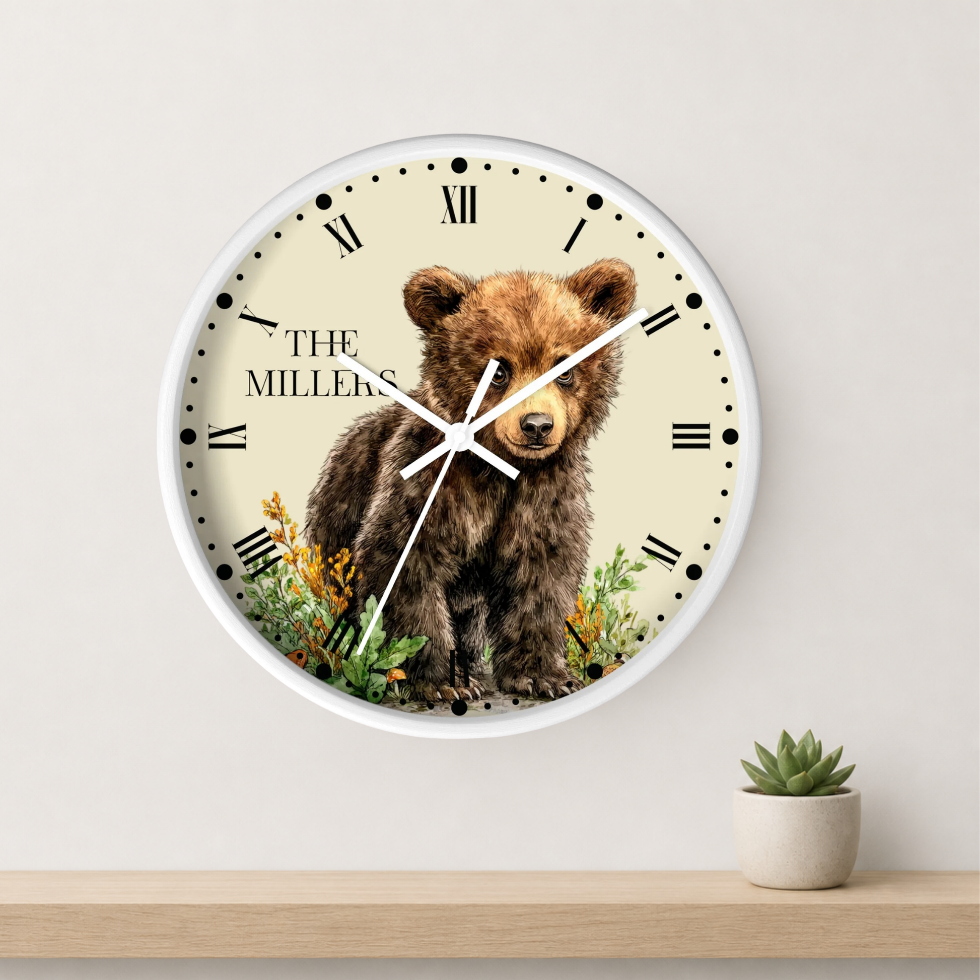 A round wall clock with a white frame and a cream dial featuring a brown bear illustration and the text 'THE MILLIONS'. The clock is mounted on a light wall above a wooden shelf with a small potted succulent.