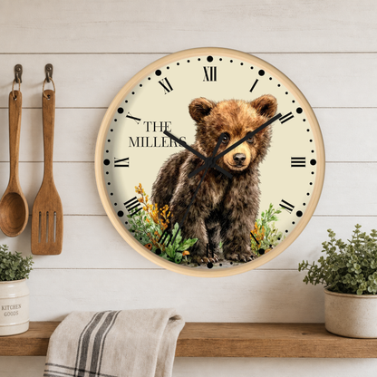 A round wall clock with a light wood frame hangs on a white wood plank wall above a shelf. The clock face features a brown bear illustration with the text 'THE MILLERS' and black Roman numerals.
