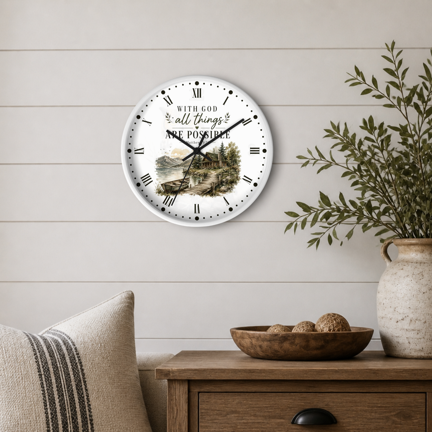 A round wall clock with a white face, black Roman numerals, and a scenic landscape illustration in the center, mounted on a light beige shiplap wall above a wooden side table with a plant and decorative bowl.