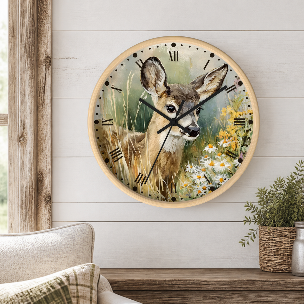 A round wall clock with a light wood frame hangs on a white shiplap wall above a wooden table. The clock face features a detailed painting of a deer in a grassy field with wildflowers.