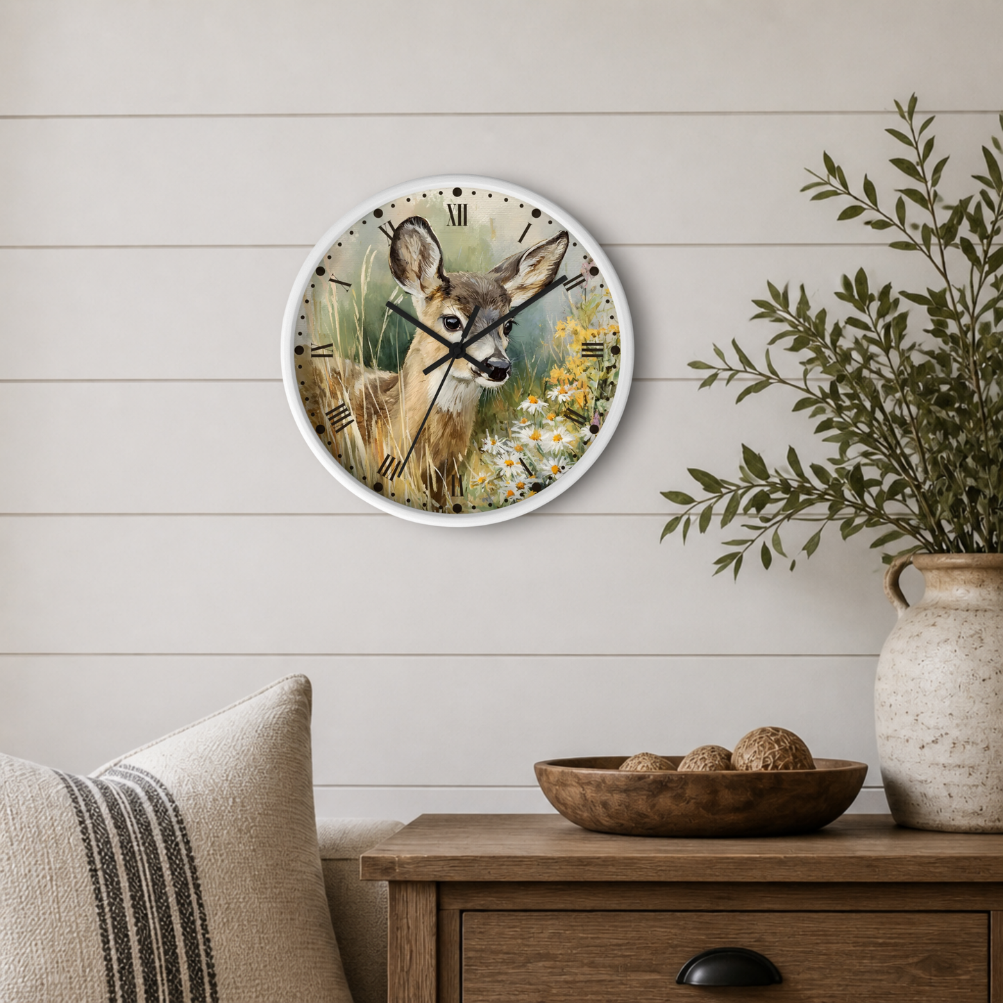 A round wall clock with a white frame hangs on a light beige shiplap wall above a wooden side table. The clock face features a painted deer in a grassy field with yellow flowers.