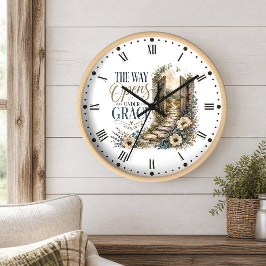 A round wall clock with a light wood frame hangs on a white shiplap wall above a wooden table. The clock face is white with black Roman numerals, black hands, and a central illustration of a stone path leading to a glowing doorway, surrounded by the words 'THE WAY Opens UNDER GRACE'.