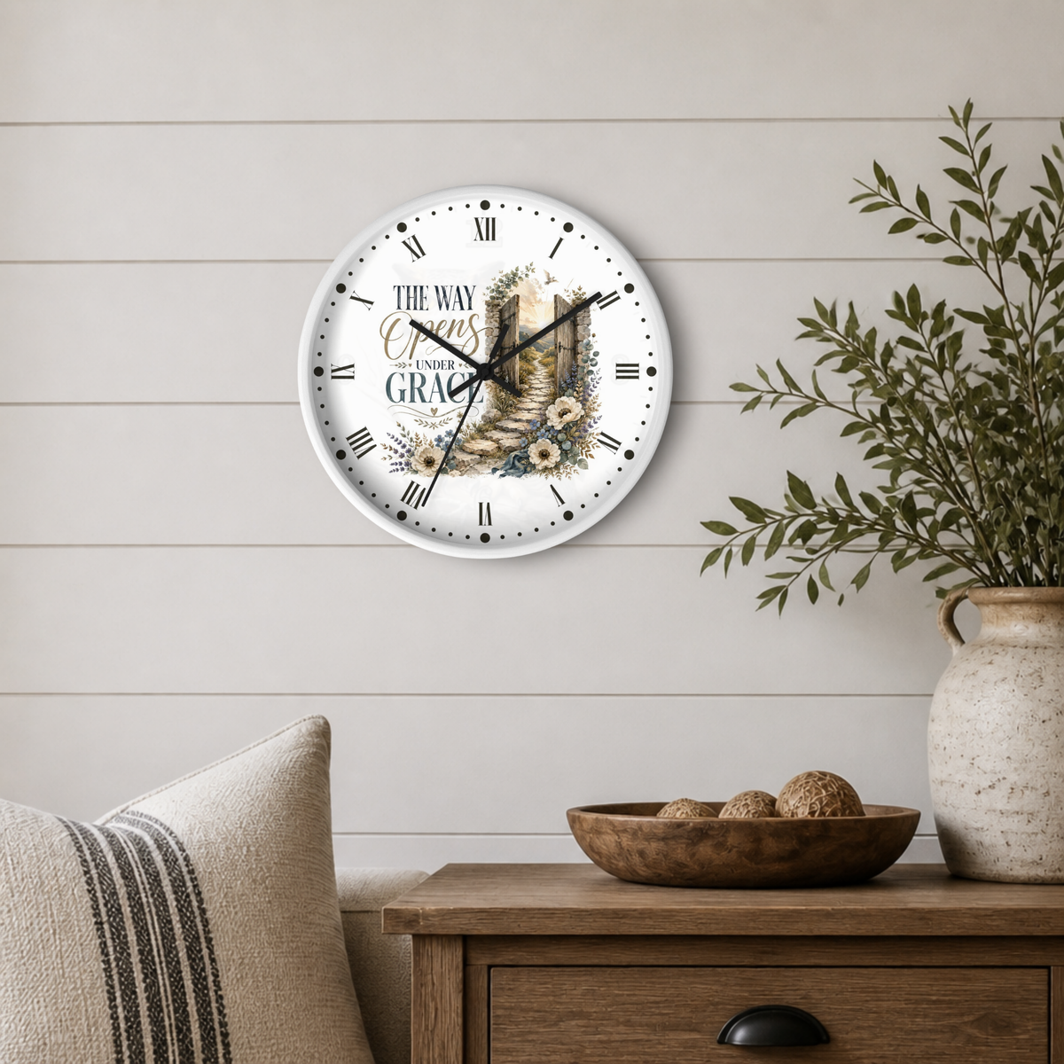 A round wall clock with a white face and black Roman numerals, featuring the text 'THE WAY OF CROSS TO GRACE' and an illustration of a stone pathway, displayed on a light wood shiplap wall above a wooden side table with a plant and decorative bowl.