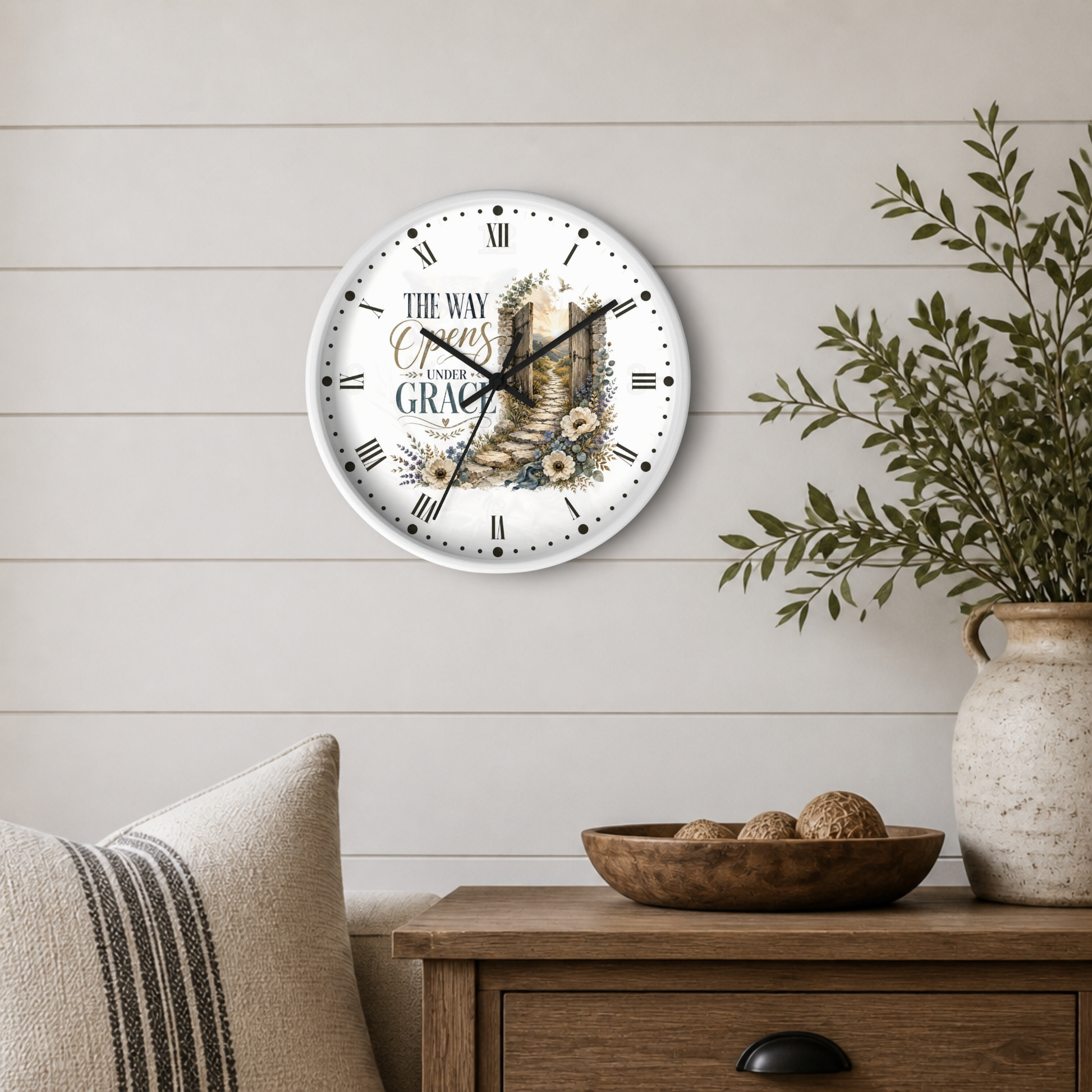 A round wall clock with a white face and black Roman numerals, featuring the text 'THE WAY OF CROSS TO GRACE' and an illustration of a stone pathway, displayed on a light wood shiplap wall above a wooden side table with a plant and decorative bowl.