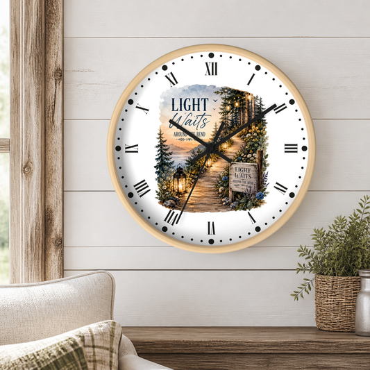 A round wall clock with a light wood frame hangs on a white shiplap wall above a wooden table. The clock face is white with black Roman numerals and hands, featuring a scenic illustration of a forest path at dusk with lanterns and the words 'LIGHT Waits Around The Bend'.