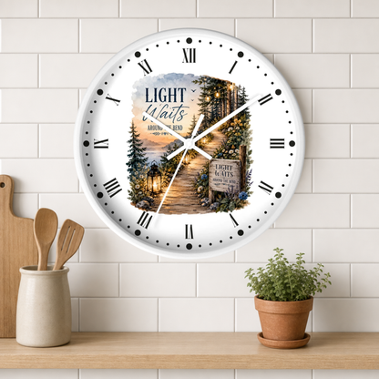 A round wall clock with a white face and black Roman numerals, featuring a scenic forest path illustration with the words 'Light Waits Around The Bend'. The clock is mounted on a white subway tile wall above a wooden shelf with kitchen utensils and a potted plant.