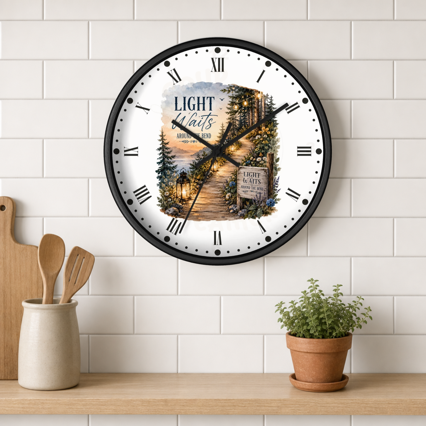 A round wall clock with a black frame and a scenic clock face illustration, mounted on a white subway tile wall above a wooden shelf with kitchen utensils and a potted plant.