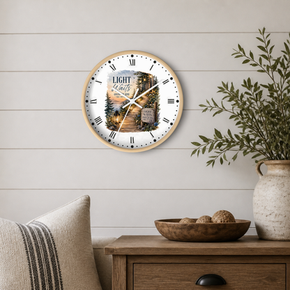 A round wall clock with a light wood frame hangs on a white shiplap wall above a wooden side table. The clock face features a scenic forest landscape with a lantern and the text 'Light waits around the bend'.