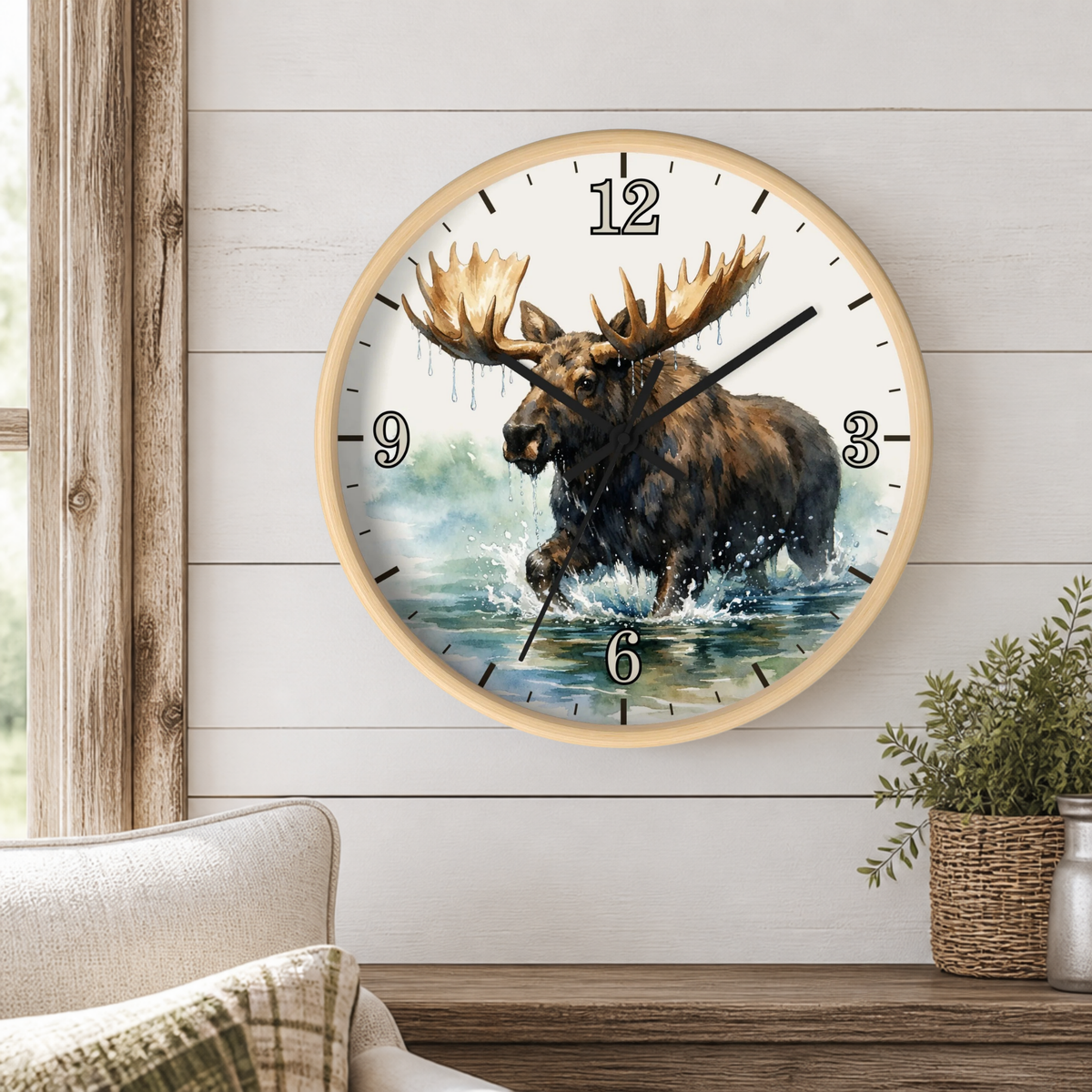 A round wall clock with a light wood frame hangs on a white shiplap wall above a wooden mantel. The clock face features a detailed illustration of a moose wading through water, with black Arabic numerals and black clock hands.